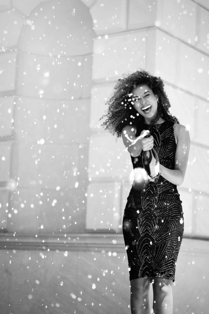 Black-and-white graduation celebration portrait of Claire Ofotokun (COFO) spraying a bottle beside pale stone architecture.