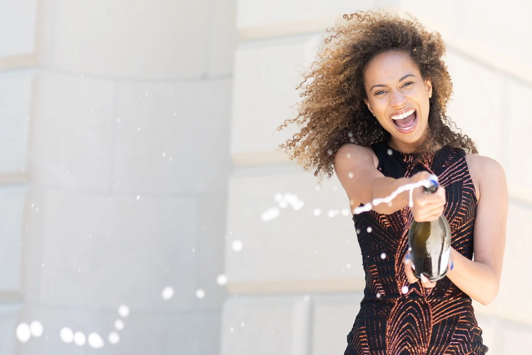 Wide color portrait of Claire Ofotokun (COFO) celebrating with a bottle spray against pale stone architecture.
