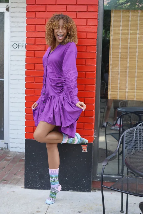 Claire Ofotokun (COFO) posing in a purple dress and colorful patterned socks beside a red brick storefront.